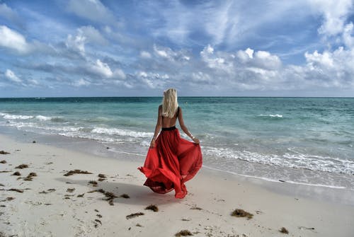 女人穿着红色的裙子，走在海边，在蓝色和白色的天空下 · 免费素材图片