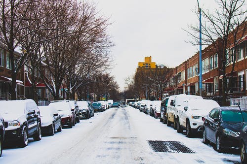 积雪覆盖的道路和白色天空下两层建筑物之间的直列停放的车辆 · 免费素材图片