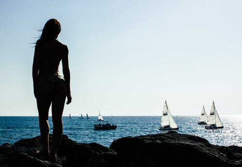 女人站在岩石与海上帆船 · 免费素材图片