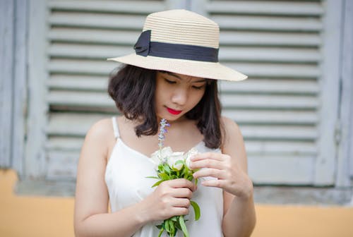 女人穿着米色太阳帽和白色无袖上衣，捧着白花 · 免费素材图片