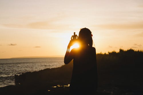 女人站在日落时附近水体的剪影 · 免费素材图片