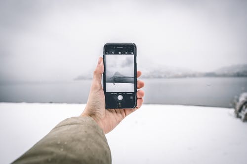 穿着棕色外套的人拿着android智能手机，同时拍摄山和水体的照片 · 免费素材图片