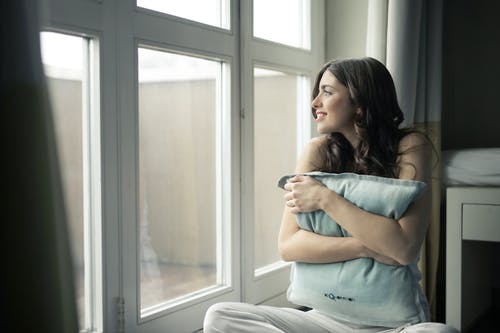 黑头发的女人在玻璃窗附近拥抱灰色枕头 · 免费素材图片