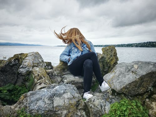 蓝色牛仔衣的女人领扣的外套和黑色的牛仔裤，坐在灰色的岩石上看湖景 · 免费素材图片