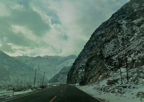 多云的天空下沥青路面附近的白雪覆盖的山脉 · 免费素材图片