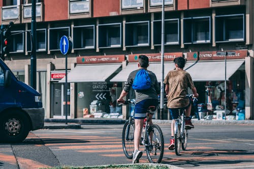 男子骑自行车在红色和灰色的大楼附近的道路上行驶 · 免费素材图片