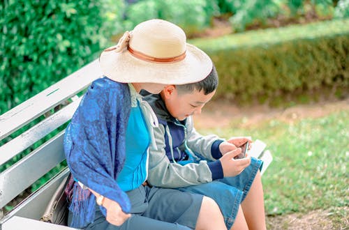 男孩身穿灰色，蓝色和黑色拉链夹克，将智能手机紧贴在旁边，身穿米色遮阳帽的人都坐在灰色的木凳上 · 免费素材图片