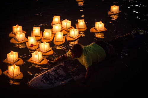 夜间，男子骑在水上纸灯笼附近的冲浪板上 · 免费素材图片