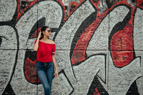 在红色和灰色的涂鸦墙附近穿红色的露肩衬衫和蓝色牛仔石洗牛仔裤的女人 · 免费素材图片