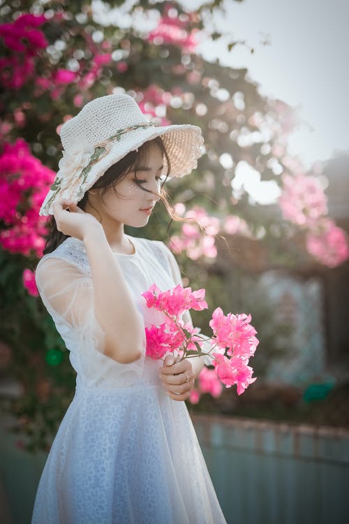 女人穿着太阳帽和持有粉红色九重葛的白色连衣裙 · 免费素材图片
