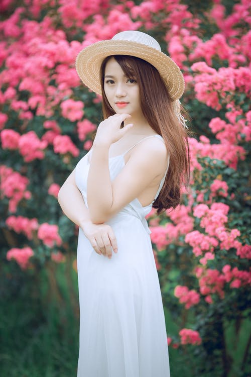 穿着白色连衣裙和遮阳帽在粉红色的花瓣花朵前的女人的景深摄影 · 免费素材图片