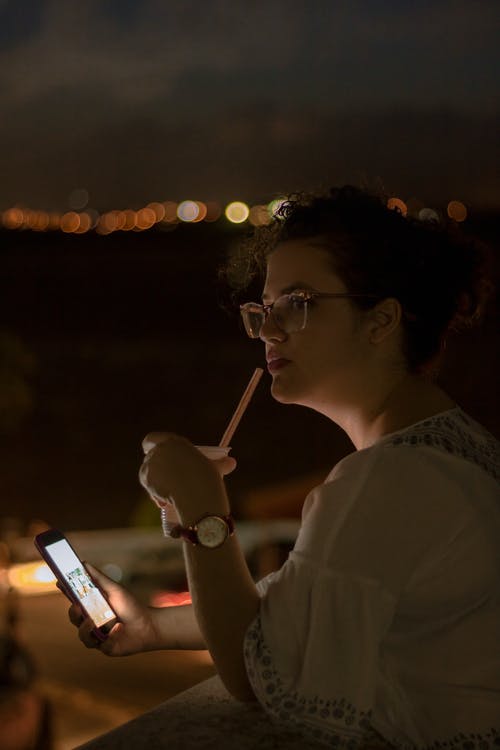 女人在夜间使用智能手机和户外举行带秸秆的塑料杯 · 免费素材图片