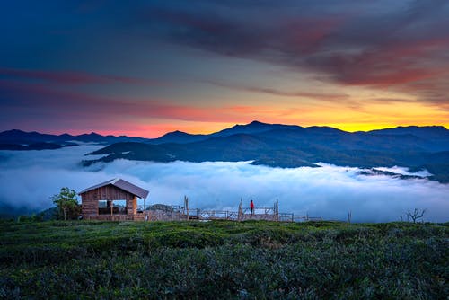 棕色木屋附近绿色的田野和橙色和黄色的天空下的山景 · 免费素材图片