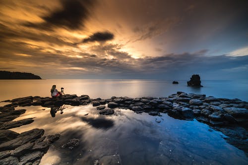 在日落时面对风平浪静的人坐在石头上 · 免费素材图片