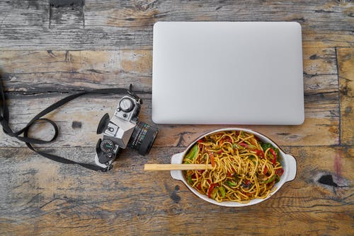 笔记本电脑和桌上的碗里的炒面旁边的相机 · 免费素材图片
