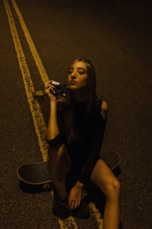 女人闭着眼睛坐在相机坐在路中间的滑板上的照片 · 免费素材图片