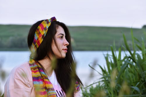 女人坐在高高的草丛与背景中的水旁边的粉红色上衣和多彩色的头巾的女人的侧视图照片 · 免费素材图片