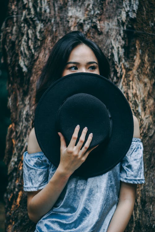 女人站在一棵树，戴着一顶黑帽子附近 · 免费素材图片