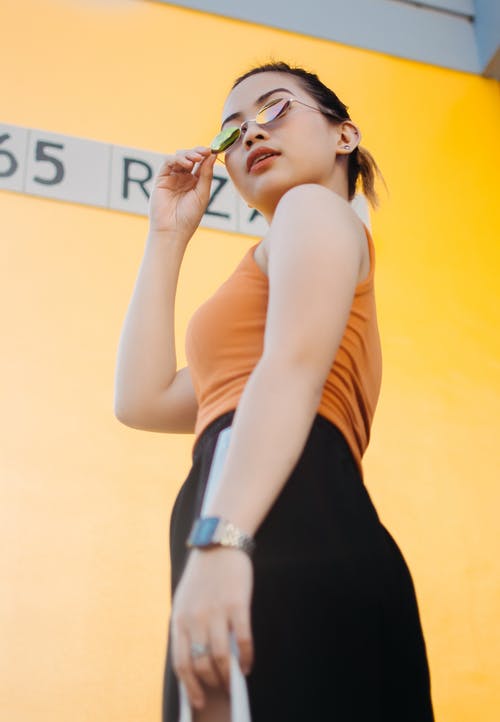 戴墨镜，橙色上衣和黑色的底部，站在黄墙旁边的女人的低角度照片 · 免费素材图片