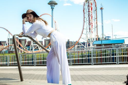 穿白色衣服的女人的照片向前倾斜在金属栏杆与游乐园在背景中 · 免费素材图片