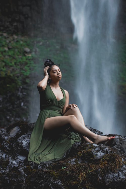 女人穿着绿色的衣服坐在岩层与瀑布在背景中的照片 · 免费素材图片
