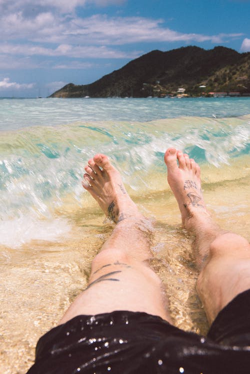 纹身的腿和一个人的脚的特写镜头，一个人躺在海边，海浪溅入岸边 · 免费素材图片