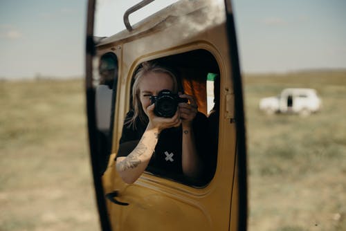 女人穿着黑色和白色的圆领t恤坐在车内同时在绿色的原野上拍照 · 免费素材图片