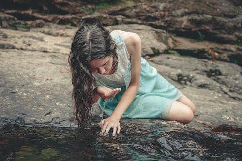 喝水时坐在水边旁边的蓝色和白色无袖连衣裙的女人 · 免费素材图片