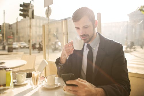 穿着黑西装的男人在一家咖啡馆喝咖啡时使用他的电话的照片 · 免费素材图片