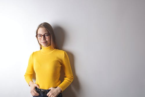 黄色的高领毛衣，身穿黑框眼镜在白墙前微笑的女人的照片 · 免费素材图片