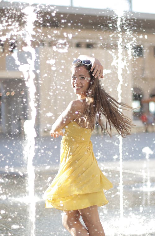 黄色的连衣裙，微笑着站在喷泉附近的女人 · 免费素材图片