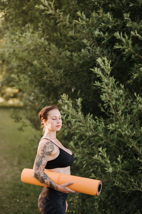 在绿色的树旁思考瑜伽垫的纹身的女人 · 免费素材图片