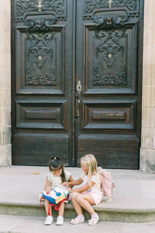 2个女孩坐在棕色木门旁边的地板上 · 免费素材图片