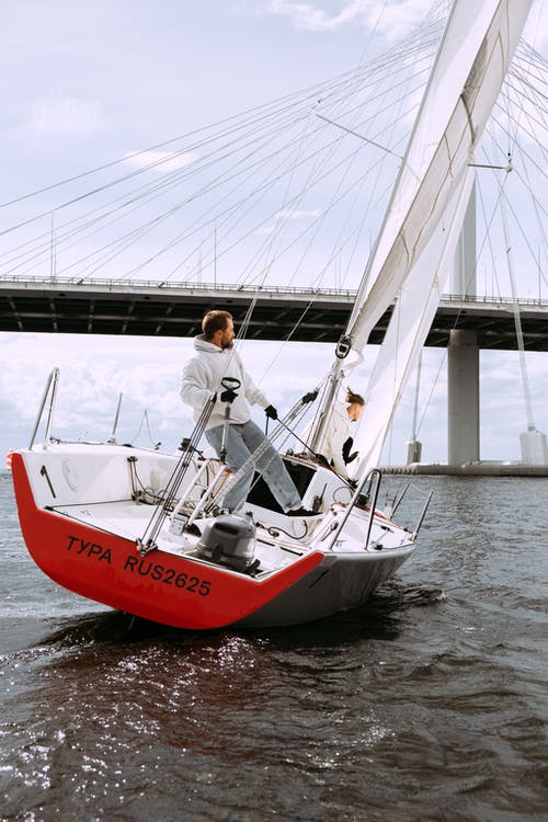 站在红色和白色的小船在水面上的白色礼服衬衫的男人 · 免费素材图片