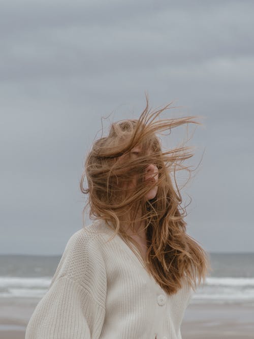 梦幻般的年轻女子，在多云的天空对海滨重新创建有风的头发 · 免费素材图片