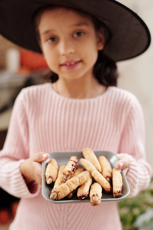 一个戴着一顶巫婆帽子和一盘巫婆手指面包设计的女孩 · 免费素材图片