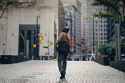 黑人男子在摩天大楼附近的城市街道上行走，看着别处 · 免费素材图片