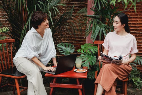 有膝上型计算机和杂志的微笑的亚裔女朋友谈话在庭院里 · 免费素材图片