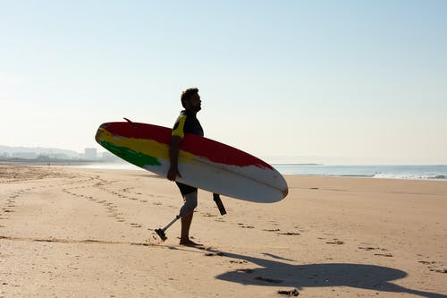 被截肢者男性冲浪者与冲浪板在海滩上行走的剪影 · 免费素材图片