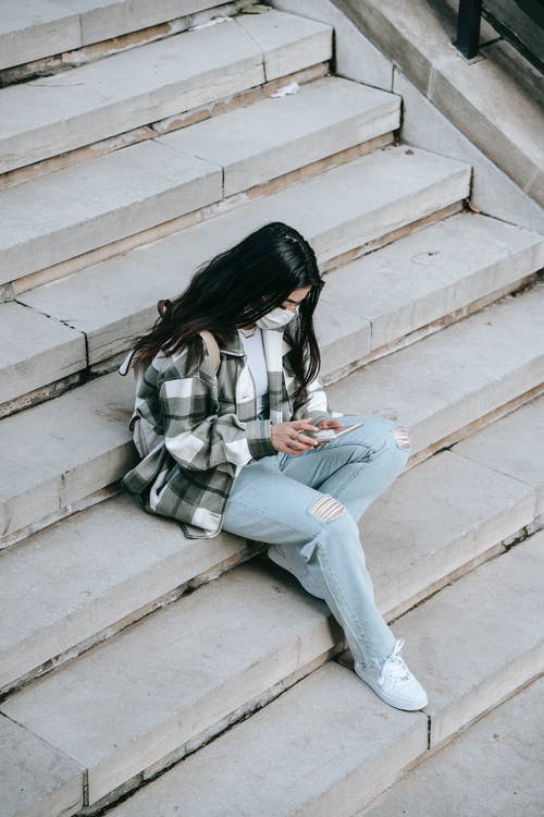 白色和黑色格子长袖衬衫和蓝色牛仔牛仔裤，坐在混凝土上的女人 · 免费素材图片