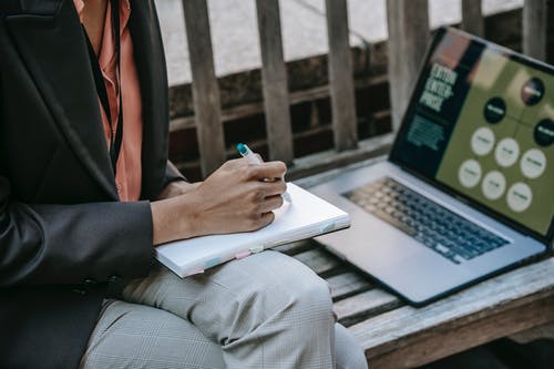 使用macbook Pro的黑色西装外套和棕色的裤子坐在长凳上的人 · 免费素材图片