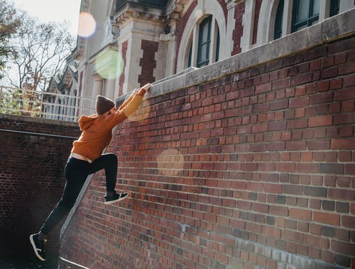 棕色的衬衫和黑色的裤子，在棕色的砖墙上跳跃的人 · 免费素材图片