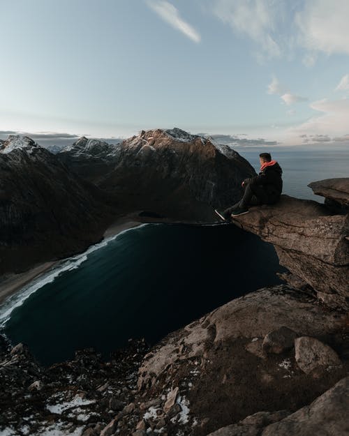 穿黑夹克的男人坐在水体附近的岩层上 · 免费素材图片