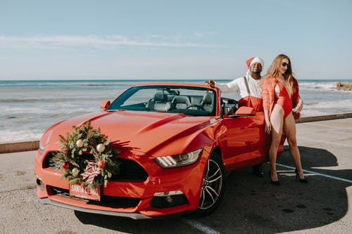 圣诞老人衣服的男人和性感的女人站在红色的车旁边 · 免费素材图片