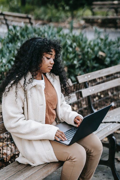 白色外套坐在长椅上使用笔记本电脑的女人 · 免费素材图片