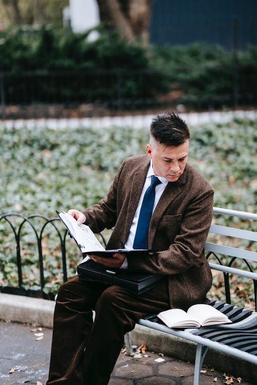 穿棕色西服外套和黑色的裤子，坐在长椅上看书的人 · 免费素材图片