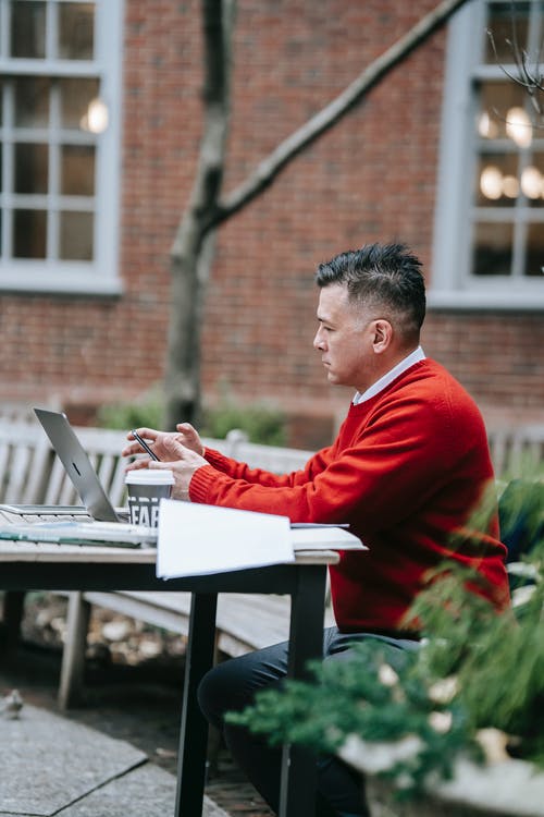红色毛衣的人在棕色的木桌上使用macbook空气 · 免费素材图片