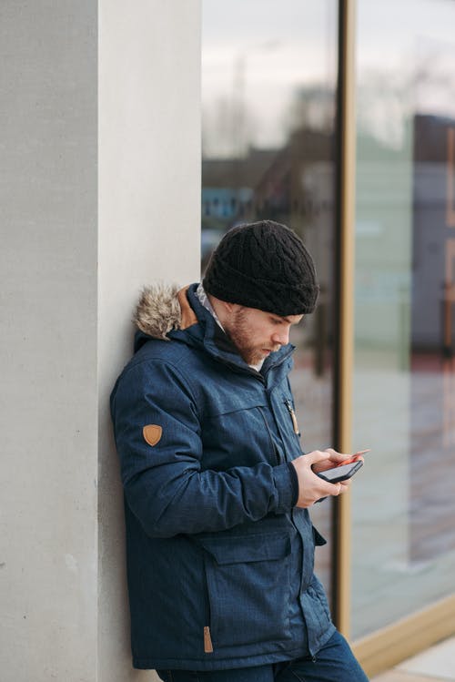 穿着蓝色夹克和黑色针织帽使用智能手机的人 · 免费素材图片