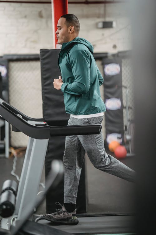 绿色外套和灰色裤子站在黑色的跑步机上的男人 · 免费素材图片