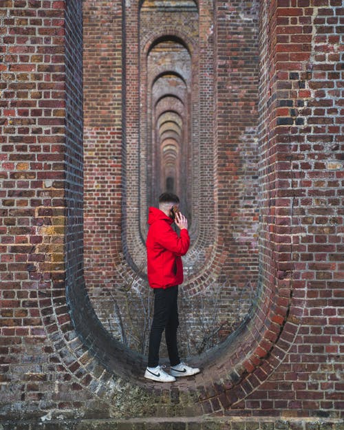 红色外套和黑裤子站立在红砖墙壁上的人 · 免费素材图片